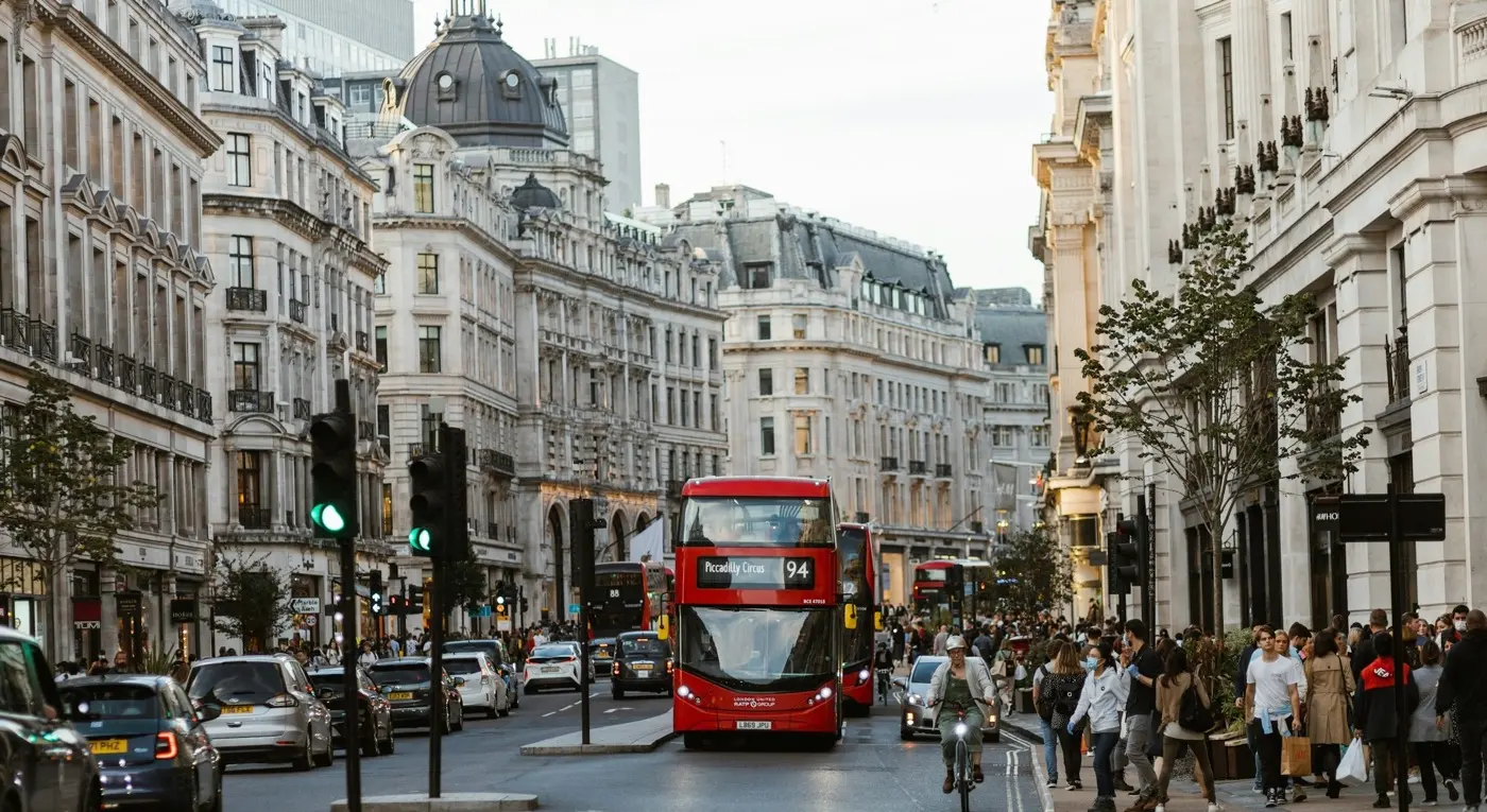 A red double-decker bus on a busy London street, stone buildings either side
